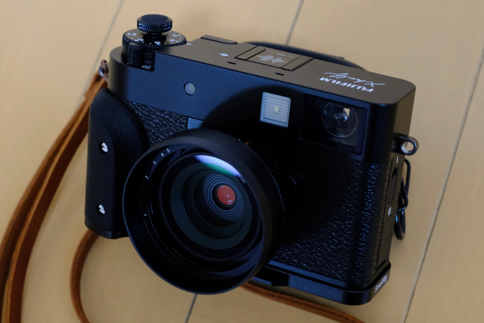 Fujifilm X half noir photographié de trois quarts sur une table en bois, avec sa sangle, une poignée et un pare-soleil tiers.
