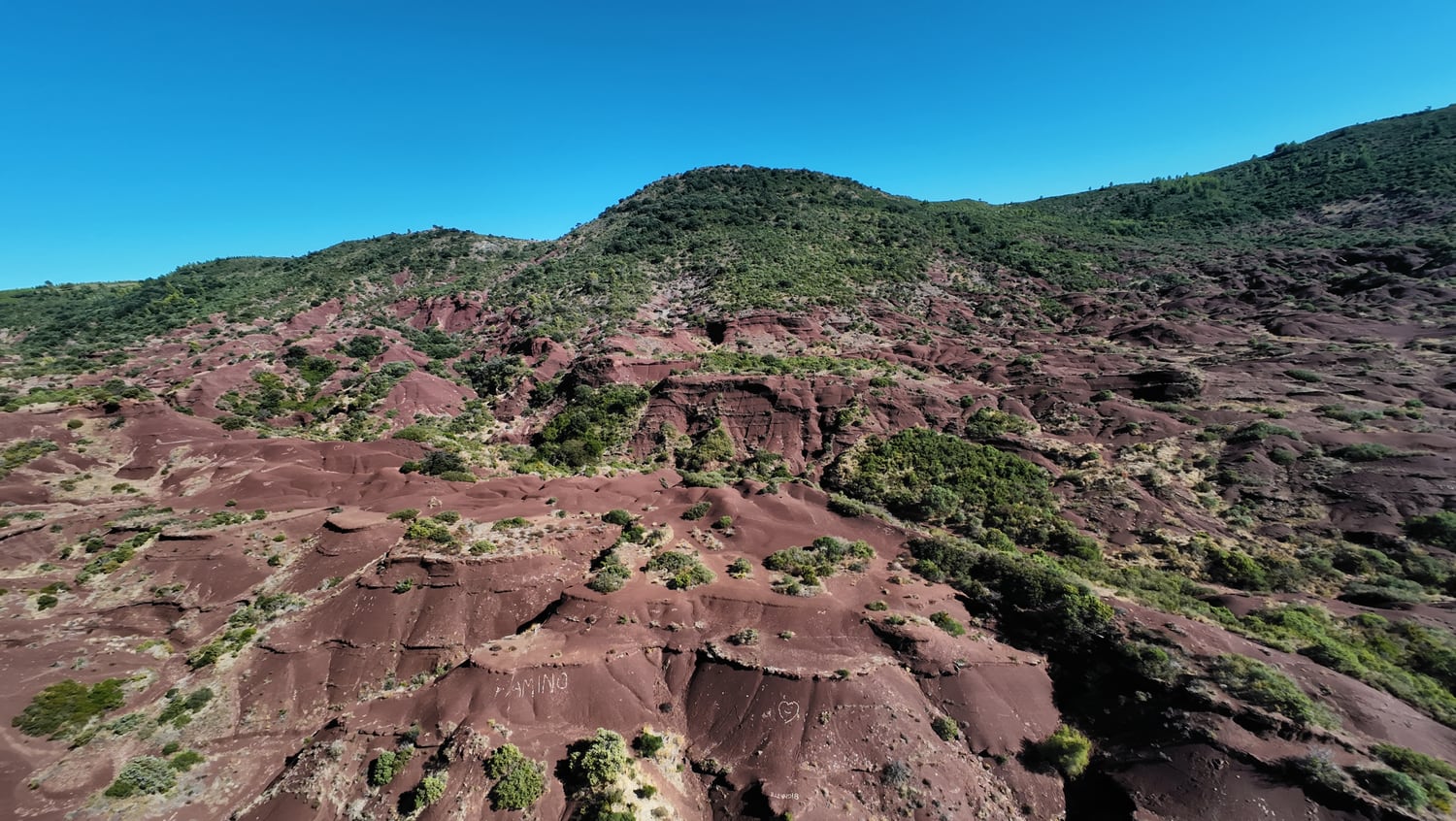 Versants rouges découpés en gradins, avec des ravines ombragées et des arbustes devant une colline verte.
