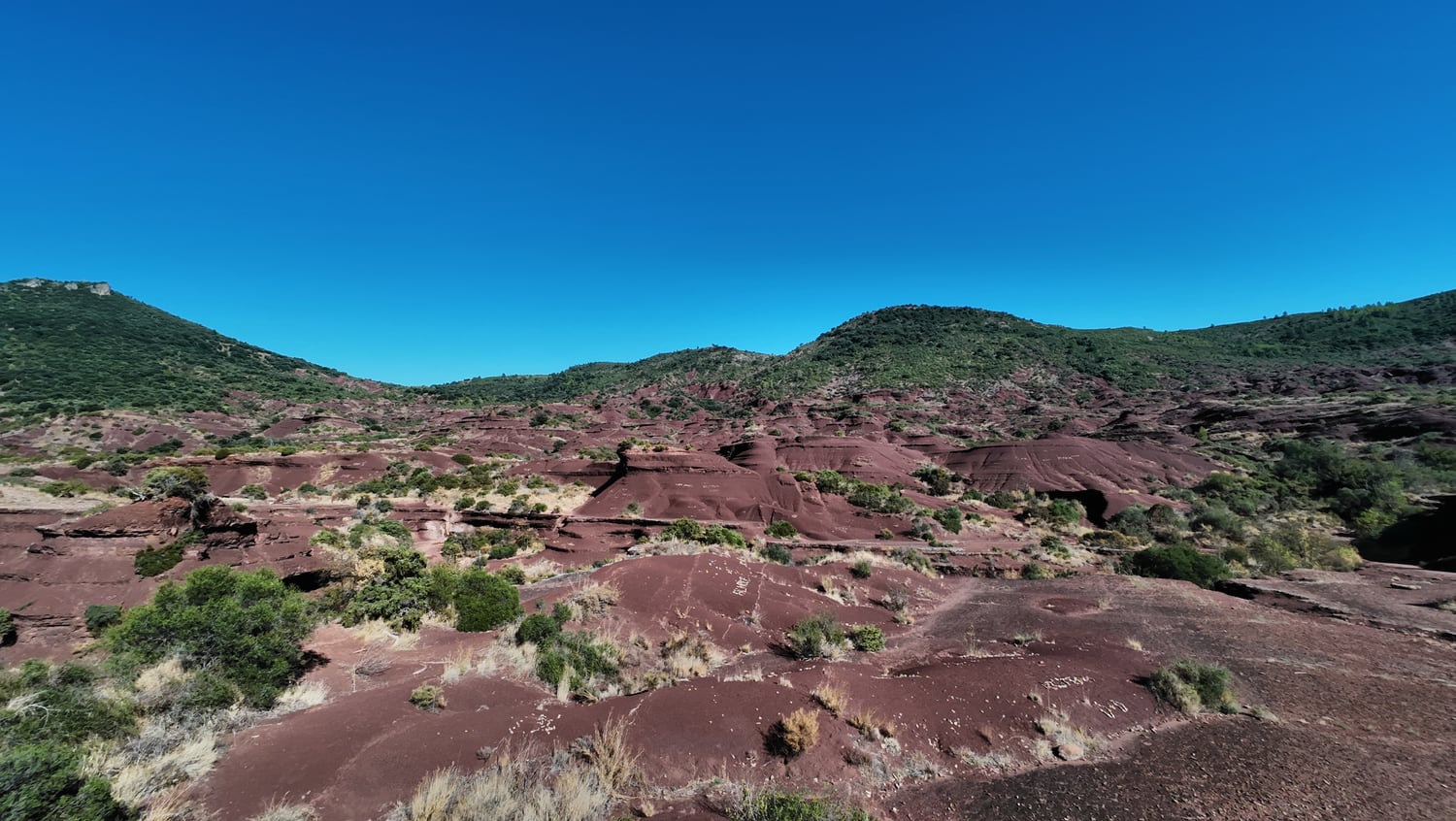 Butte rouge au centre du Canyon du Diable, entourée de surfaces ravinées, d'arbustes et de collines sous un ciel bleu.