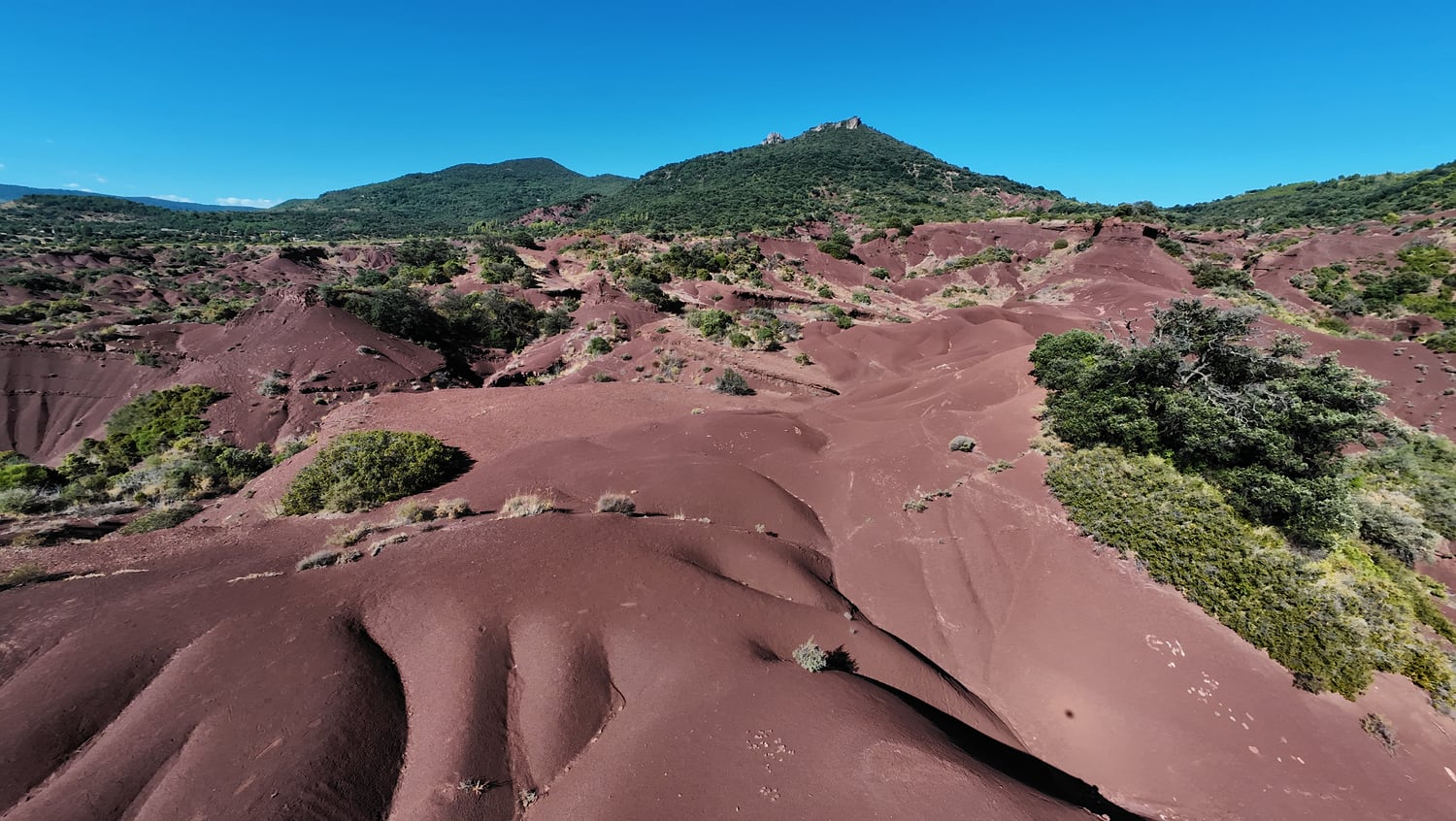 Reliefs rouges ravinés du Canyon du Diable, avec des arbustes et une colline boisée en arrière-plan, vus depuis le DJI Avata 2.