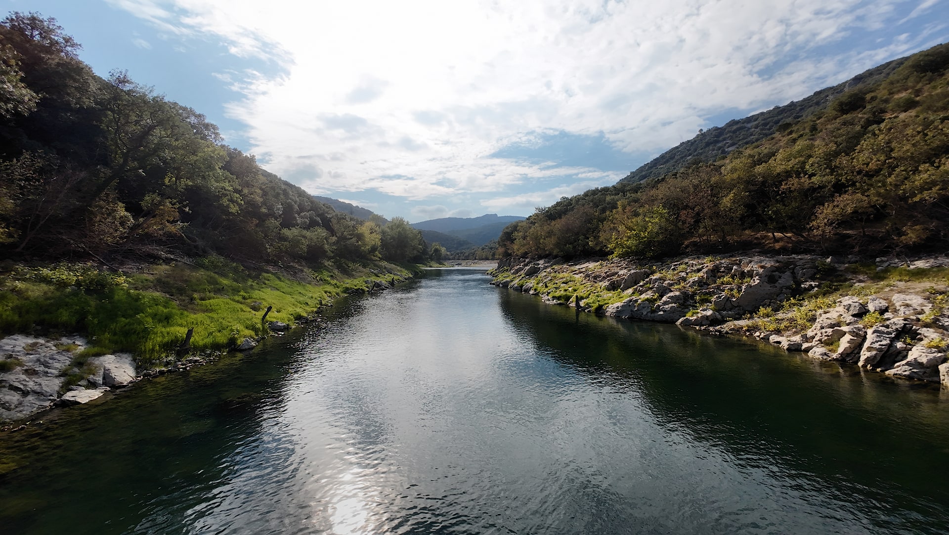 Vue proche de la surface de l'Hérault, avec les reflets du ciel entre des berges rocheuses et boisées.