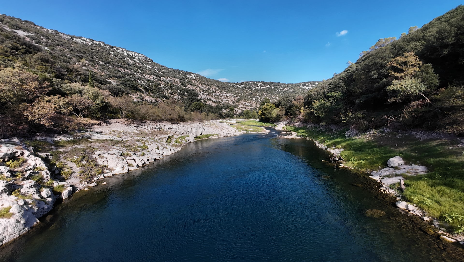 Eau sombre de l'Hérault sous un ciel bleu, entre une rive rocheuse claire à gauche et un versant boisé à droite.