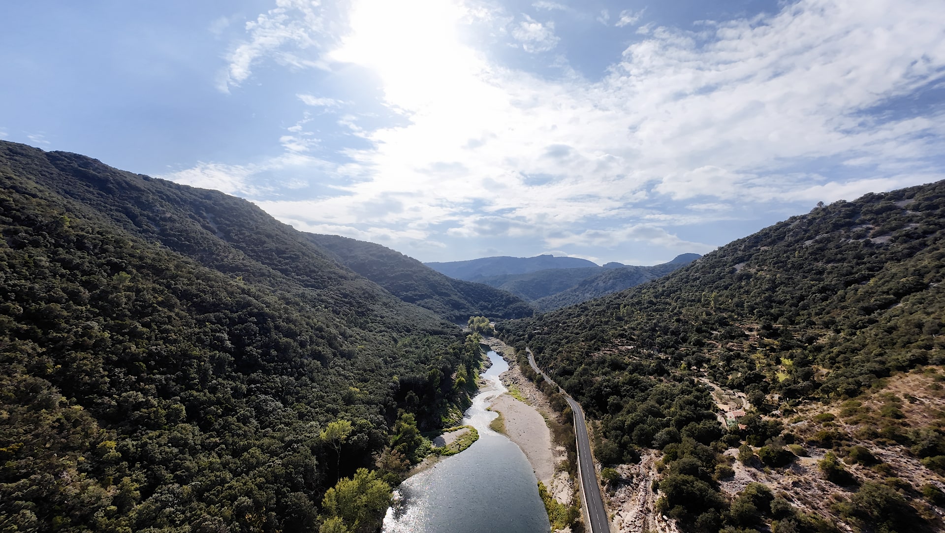 L'Hérault réfléchissant la lumière entre deux versants boisés, avec des collines successives à l'horizon et une route sur la droite.
