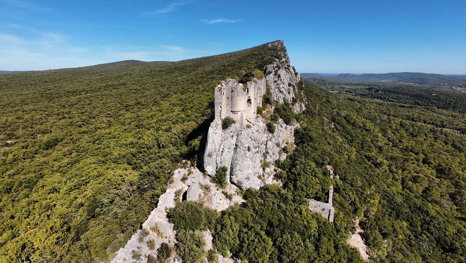Ruines de Viviourès vues de biais, avec une haute façade ajourée et des murs suivant le bord du rocher.