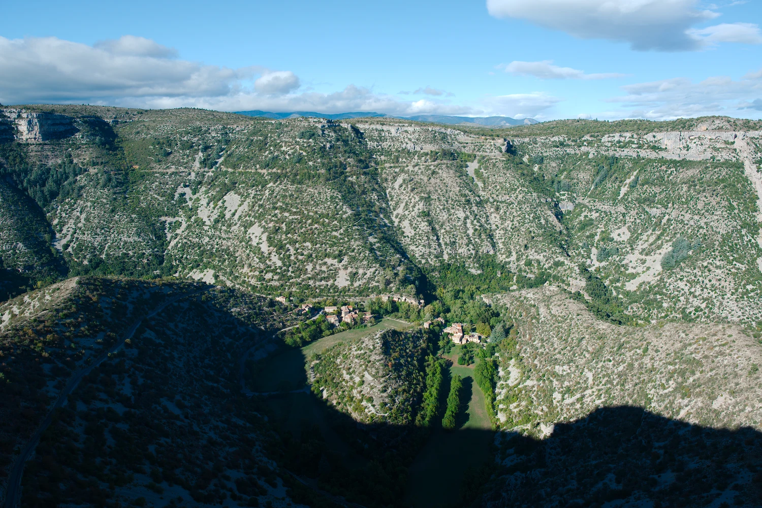 Le méandre de Navacelles et ses maisons sous les falaises, entre une zone éclairée et l’ombre du versant au premier plan.