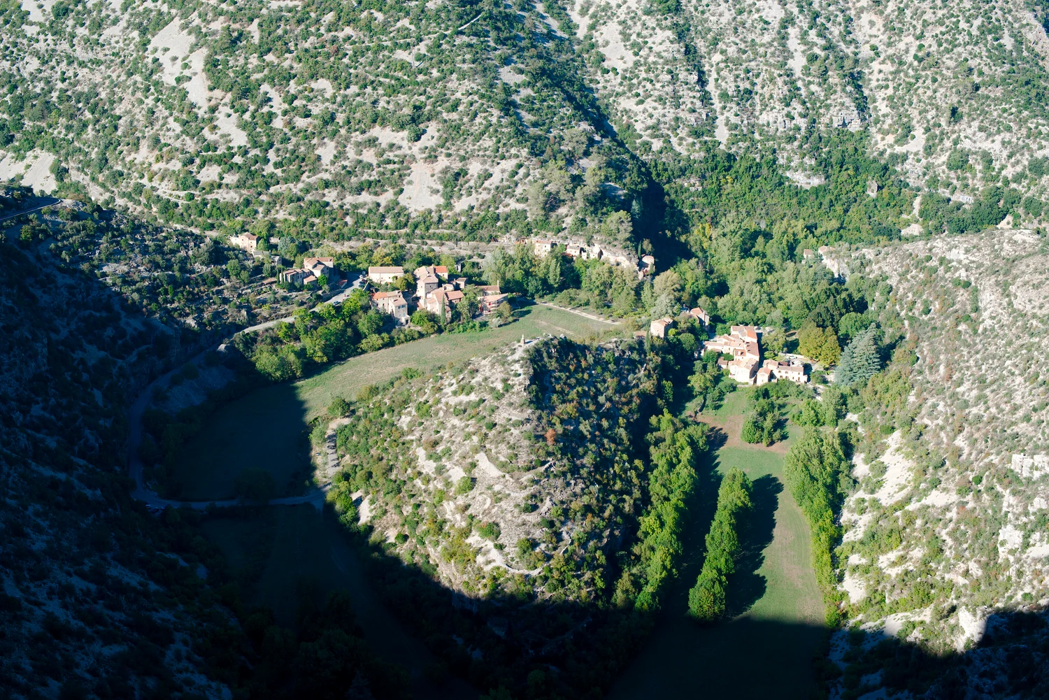 Vue rapprochée des maisons et de la prairie courbe autour du relief rocheux central de Navacelles, partiellement dans l’ombre.