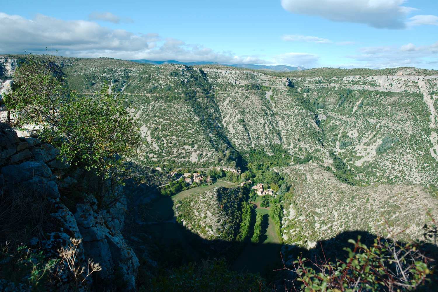 Roches et arbustes au premier plan à gauche du cirque de Navacelles, avec le méandre et les maisons plus bas.