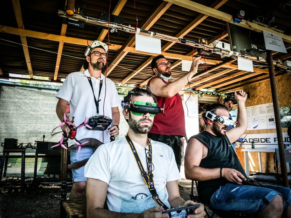 Des pilotes italiens réunis sous un abri ouvert, avec radiocommandes, lunettes FPV et un petit drone.